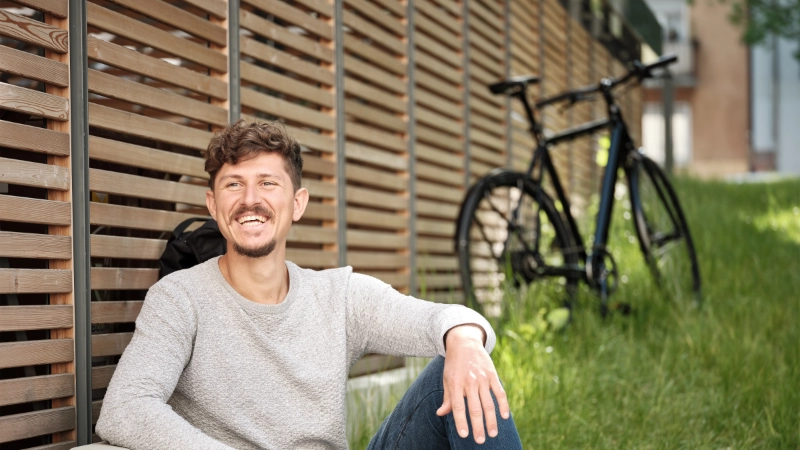Ein Fahrradfahrer lehnt an einen Holzzaun und lacht. Sein schwarzes Fahrrad steht neben ihm, an den Zaun angelehnt.